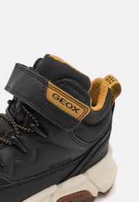 Černá a hořčicová teniska Geox s páskem na suchý zip, polstrovaným límcem, šněrováním a masivní bílou podrážkou, zobrazená zblízka.