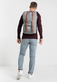 Sac à dos gris avec des sangles et des accents en cuir marron, design à rabat, et une poche frontale extérieure. Fabriqué en tissu durable.