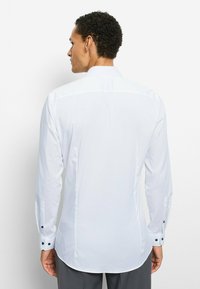 Weißes Langarmshirt mit geschwungenem Saum, geknöpften Bündchen mit marineblauen Akzenten und glatter Textur, von hinten gezeigt.