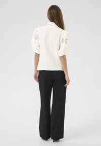 Blusa corta bianca e texturizzata con dettagli in pizzo, abbinata a pantaloni neri larghi. La blusa presenta un colletto e maniche tonde.
