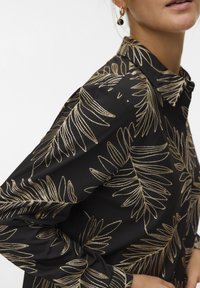 Vrouw met een zwart shirt met een metallic gouden bladpatroon, gecombineerd met gouden en zwarte oorringen, handen in de zij.