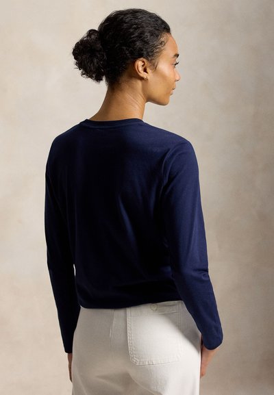 Γυναίκα με σκούρα σγουρά μαλλιά μαζεμένα σε κότσο, φοράει ένα μπλε μακρυμάνικο τοπ και λευκό παντελόνι, βλέποντας προς τα πίσω σε ουδέτερο φόντο.