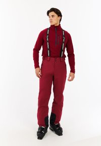 Rode zip-up jas met een voorkraak, gecombineerd met rode skibroeken met zwarte onderaccenten en verstelbare bretels. Zwarte skibotten.