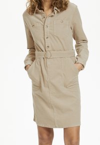 Abito camicia in corduroy beige con maniche lunghe, chiusura a bottoni, due tasche sul petto, vita con cintura e anello dorato, e tasche laterali.