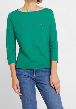 Persoon draagt een groen shirt met boothals en lange mouwen, en een blauwe spijkerbroek, staand met de ene hand ontspannen en de andere rustend op de dij.