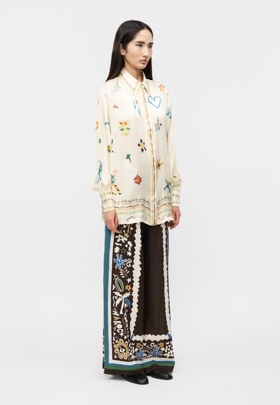 Νεαρή γυναίκα που φοράει μια φαρδιά κρεμ μπλούζα με πολύχρωμα λουλουδένια σχέδια και παντελόνι με φαρδιά πόδια, διακοσμημένο με περίπλοκα μοτίβα με μπλε και πράσινα περιθώρια.