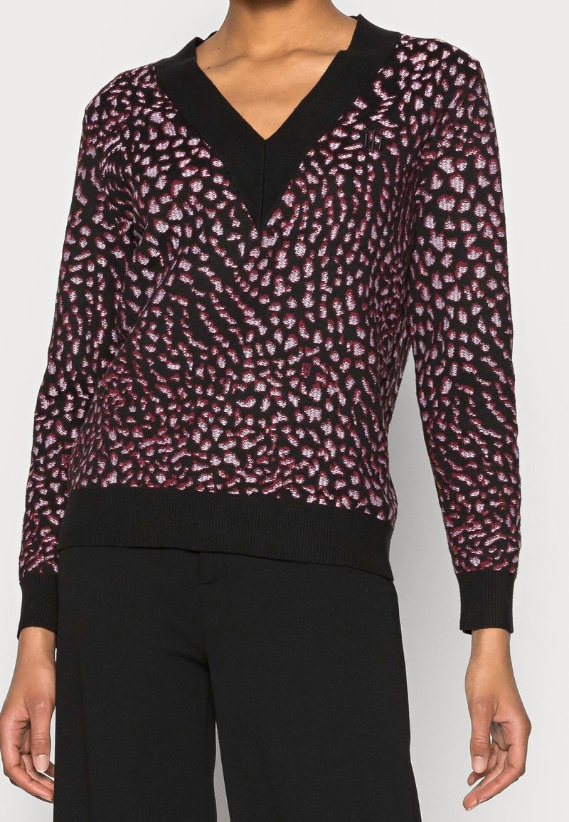 Femme portant un pull noir avec un motif abstrait rose et blanc et des poignets et un bas en côtes noirs, associé à un pantalon noir.