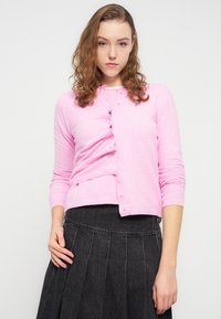 Ροζ πλεκτή ζακέτα με μακριά μανίκια, με κουμπιά μπροστά και εφαρμοστό σχέδιο, συνδυασμένη με μαύρη πλισέ φούστα.