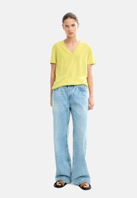 Żółta koszulka z krótkim rękawem i dekoltem w serek, zestawiona z jasnoniebieskimi, wysokim stanem dzwonami i czarnymi sandałami. Gładki materiał, luźny krój, casualowy styl.