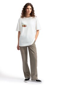 Witte katoenen t-shirt met korte mouwen, met een kleurrafische afbeelding op de voorkant. Gecombineerd met losse beige broeken en zwarte schoenen.