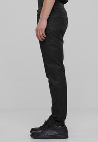 Čierne denimbové nohavice so slim fit strihom, vybavené štandardnými vreckami a hladkou textúrou. V kombinácii s čiernymi teniskami. Bočný pohľad na šedom pozadí.