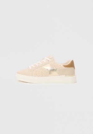 Beige Sneaker mit einem Materialmix-Design, das flauschige Akzente, metallische Details und einen strukturierten Rücken aufweist, auf einer weißen Sohle.