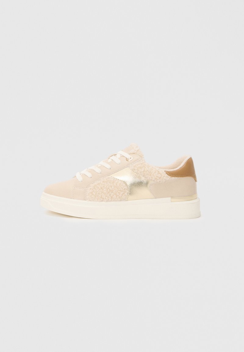 Beige sneakers with a mixed material design, featuring fluffy accents, metallic details, and a textured back, set on a white sole.