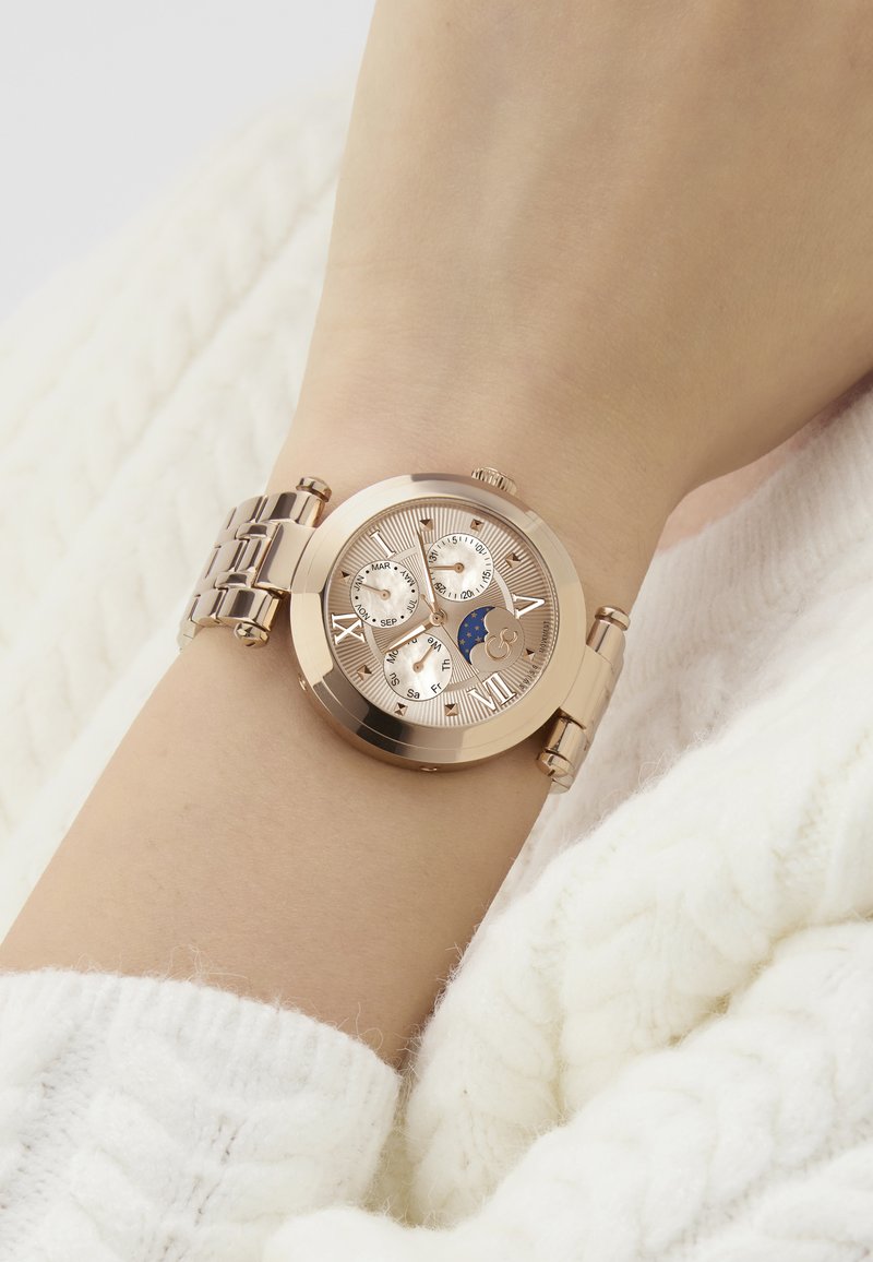 Różowe złote zegarek na nadgarstku, z fakturowaną tarczą z oznaczeniami daty, trzema subtarczami i niebieskim wskaźnikiem faz Księżyca. Metalowa bransoleta.