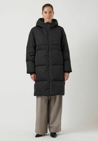 Piumino nero con cappuccio, caratterizzato da una chiusura frontale con zip, una vestibilità oversize e sezioni trapuntate. Indossato sopra pantaloni beige chiaro e scarpe nere.