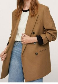 Brun dobbeltradet blazer med sorte knapper, skræddersyet pasform, blød tekstur og oprullede ærmer, båret over en hvid skjorte og blå jeans.