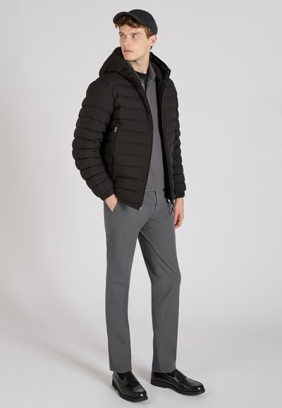 Zwarte puffy jas met capuchon, met ritszakken; gedragen over een grijs shirt en grijze broek; bijpassende zwarte schoenen. Eenvoudige achtergrond.