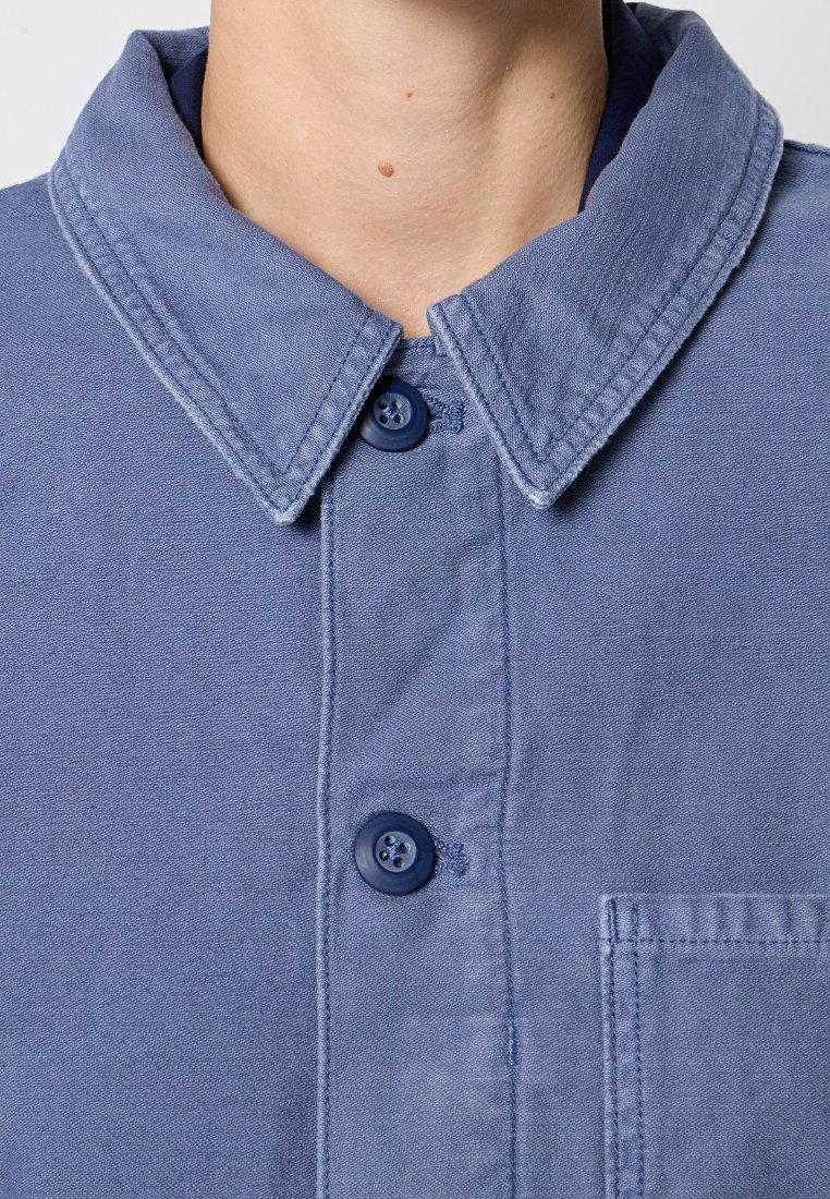 Nahaufnahme eines navyblauen, knopfartigen Hemdkragens und Oberkörpers mit sichtbaren Knöpfen und gestepptem Tasche auf heller Haut.