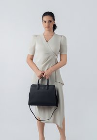 Borsa in pelle nera texturizzata con doppie maniglie e tracolla staccabile, abbinata a un vestito avvolgente color beige chiaro.