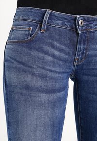 Detailní záběr na středně modré džíny s předními kapsami, poutky na pásek a kovovým knoflíkem, nošené s černým topem.