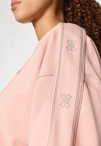 Rózsaszín pulóver, amely puha textúrával, ezüst gyöngysorral díszített vállakkal és logó mintákkal rendelkezik, valamint laza fazonú.