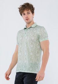 Camisa polo verde texturizada con cuello, dos botones y mangas cortas. Usada con jeans oscuros frente a un fondo liso.