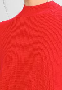 Red ribbed knit sweater featuring a high neckline and textured fabric, emphasizing a close-fitting design without visible seams or embellishments.