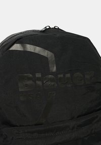 Zaino nero con una superficie testurizzata, dotato di un logo in rilievo prominente e di un design elegante e minimalista. Chiusura a zip sulla parte superiore.