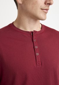 Maroon henley shirt featuring a round neckline, four button placket, and subtle ribbed texture around the collar and cuffs.