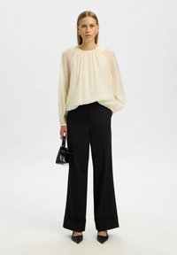 Kremet chiffonbluse med elastisk halsudskæring, sorte skræddersyede bukser med vidde i benene og spidsede sorte hæle, holder en lille sort håndtaske.