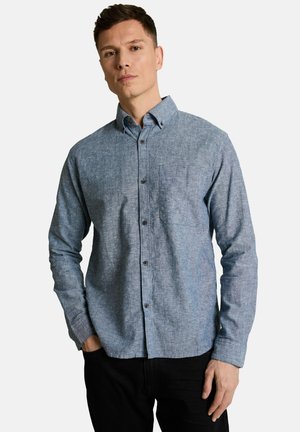 Mann mit blauem langärmeligen Hemd mit Brusttasche, stehend vor einem einfarbigen hellen Hintergrund.