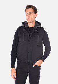 Gilet nero in tessuto testurizzato, con cappuccio grigio, due tasche frontali, chiusura con zip e coulisse regolabili. Indossato sopra una maglietta a maniche lunghe nera.