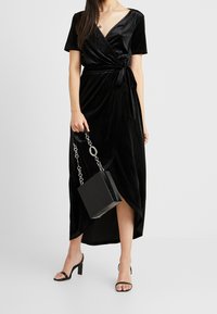 Schwarzes Samt-Wickelkleid mit kurzen Ärmeln und tailliertem Bund, kombiniert mit einer strukturierten schwarzen Handtasche und schwarzen Sandaletten.