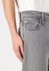 Hallid denim teksad sirge lõikega, viie tasku, metallist konksude ja sileda tekstuuriga. Suurendus vöökoha ja käte asendi kohta.