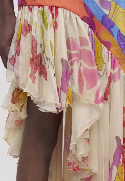Διαφανής, πολυεπίπεδη φούστα με πολύχρωμα λουλουδάτα μοτίβα και βολάν στα άκρα, που αποκαλύπτει εν μέρει σκουρόχρωμο πόδι πάνω σε ουδέτερο φόντο.