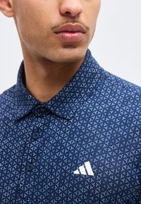 Κοντινό πλάνο ενός άνδρα που φοράει μπλε σκούρο πουκάμισο με λευκά γεωμετρικά σχέδια και ένα λευκό λογότυπο Adidas στο στήθος.