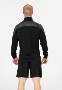 Man i svart idrottsjacka med grå mönster på axlarna och svarta shorts, stående vänd bort från kameran mot vit bakgrund.