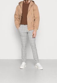 Beige huvjacka med en brun skjorta under, tillsammans med ljusgrå rutiga byxor och vita sneakers; avslappnad stil.