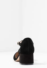 Zapato de ante negro con punta redondeada y un tacón grueso. Presenta una correa lateral con una hebilla dorada y una superficie lisa y texturizada.