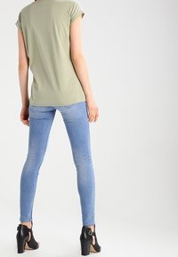 Osoba v svetle zeleném tričku s krátkým rukávem, světlých modrých skinny džínech a černých sandálech na podpatku stojí zády k jednoduchému pozadí.