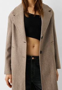 Cappotto di lana color marrone chiaro con colletto a scollo, due bottoni e tasche laterali; indossato sopra un top corto a coste nero e jeans scuri.