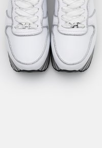 Bílé tenisky se stříbrnými akcenty, hladký kožený svršek, texturovaná oblast šněrování a gumová podrážka s černým pruhem. Obsahují detaily s metalickým logem.