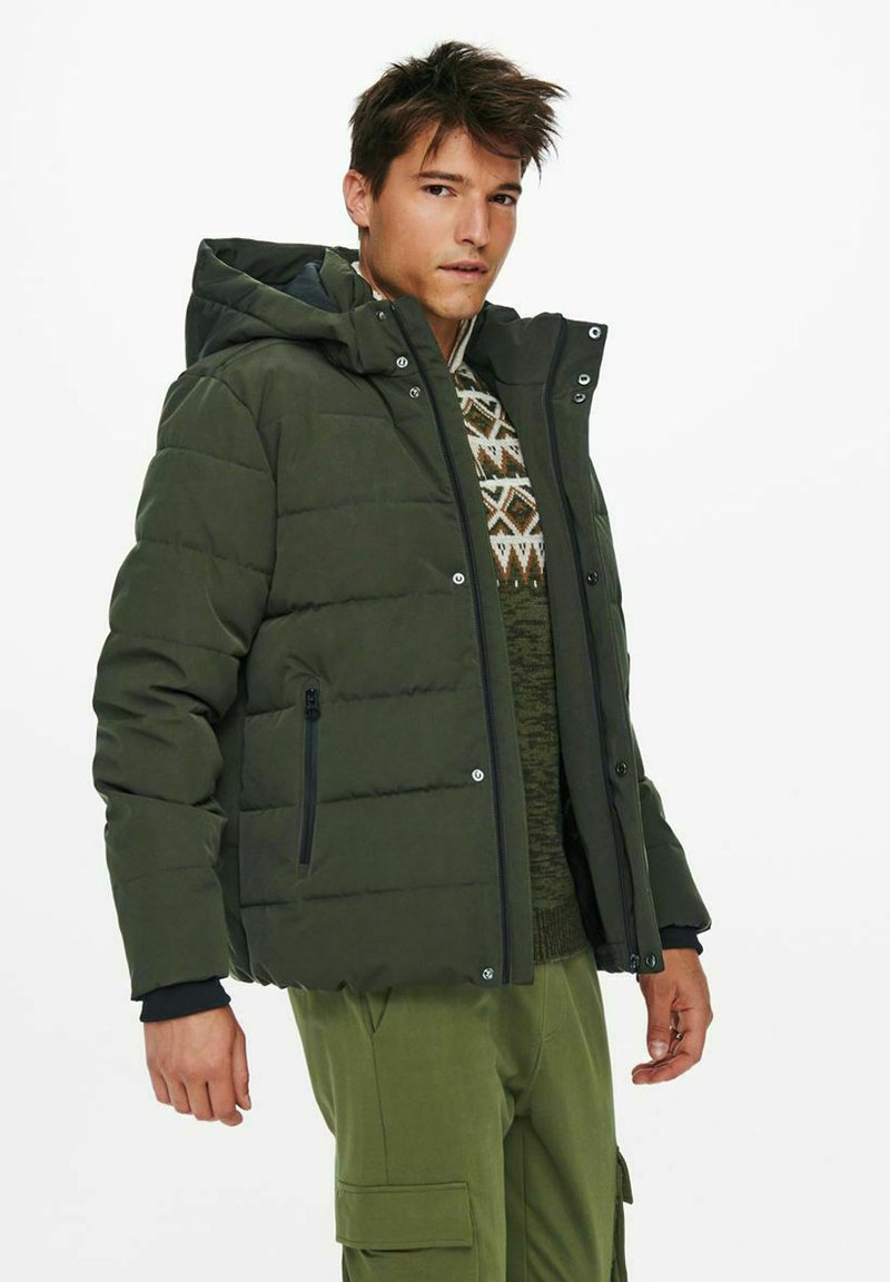 Olive green puffer jacket with a zip closure, hood, and side pockets. Features quilted texture and silver hardware accents. Casual design.