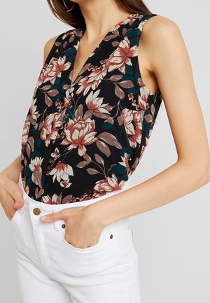 Sleeveless black top with large pink and beige floral print worn with white pants, hands tucked in front pockets.
