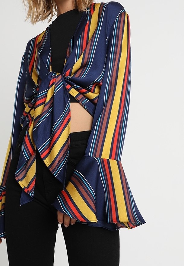 Femme portant un chemisier à rayures multicolores bleu marine avec un nœud à l'avant et des manches cloche évasées, associé à un pantalon noir.