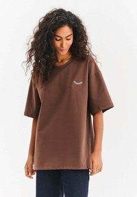 Brązowa, oversize'owa koszulka z miękkiej bawełny. Ma krótkie rękawy oraz subtelny biały wzór z kreskówek na klatce piersiowej. Casualowy krój.