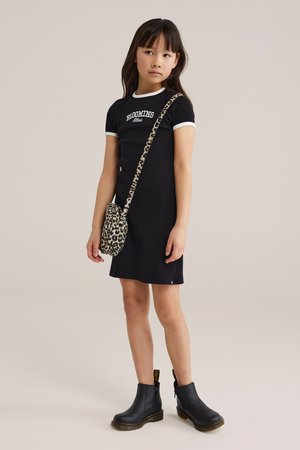 Lány fekete ruhában, amelyen a "BLOOMING" felirat szerepel, leopárdmintás oldaltáskával és fekete bokacsizmában, egyszerű háttér előtt áll.