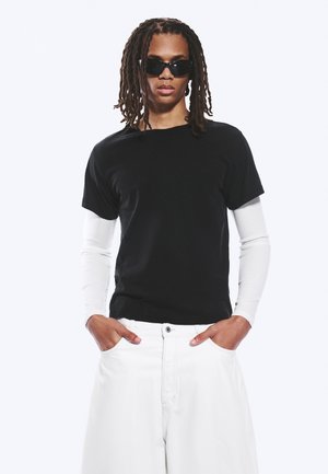 Personne avec des dreadlocks portant des lunettes de soleil noires, une chemise noire à manches courtes sur une chemise blanche à manches longues, et un pantalon blanc les mains dans les poches.