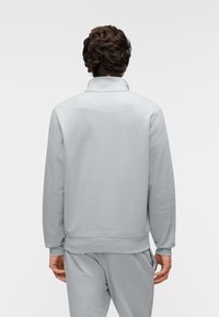 Ανοιχτό γκρι φούτερ με ψηλό γιακά, μακριά μανίκια και ριμπ τελείωμα. Λείο υλικό, μινιμαλιστικός σχεδιασμός χωρίς έντονες λεπτομέρειες ή διακοσμητικά στοιχεία.