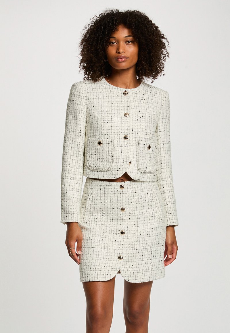 Donna che indossa un blazer bianco texture con bottoni e una gonna coordinata con tasche, in piedi contro uno sfondo chiaro e uniforme.